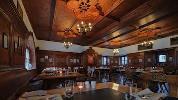 Une salle à manger spacieuse, avec des boiseries et des lustres. Les tables sont dressées avec des couverts et des verres.