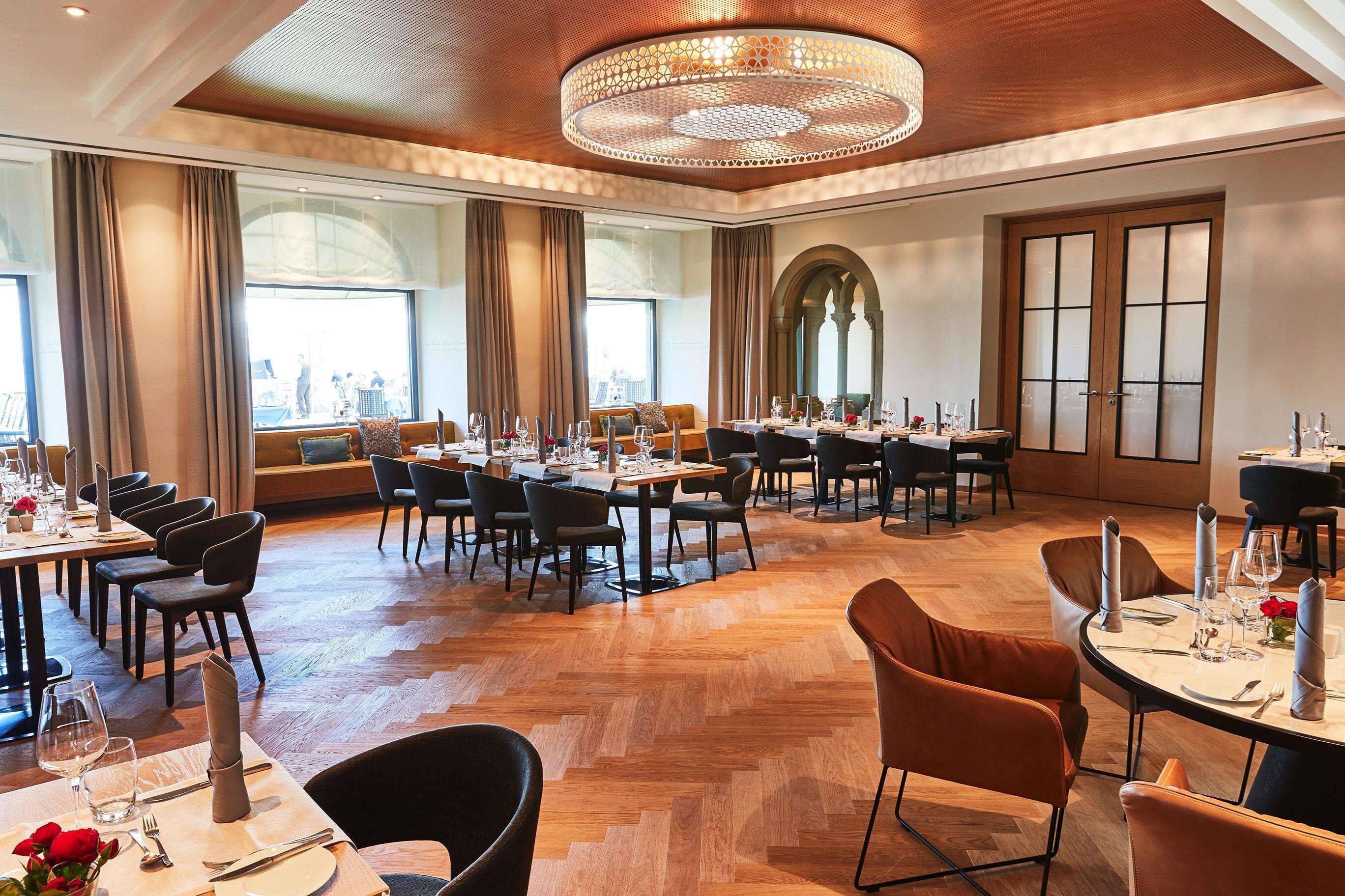 A spacious dining area with multiple tables and chairs, large windows, and a decorative ceiling light.