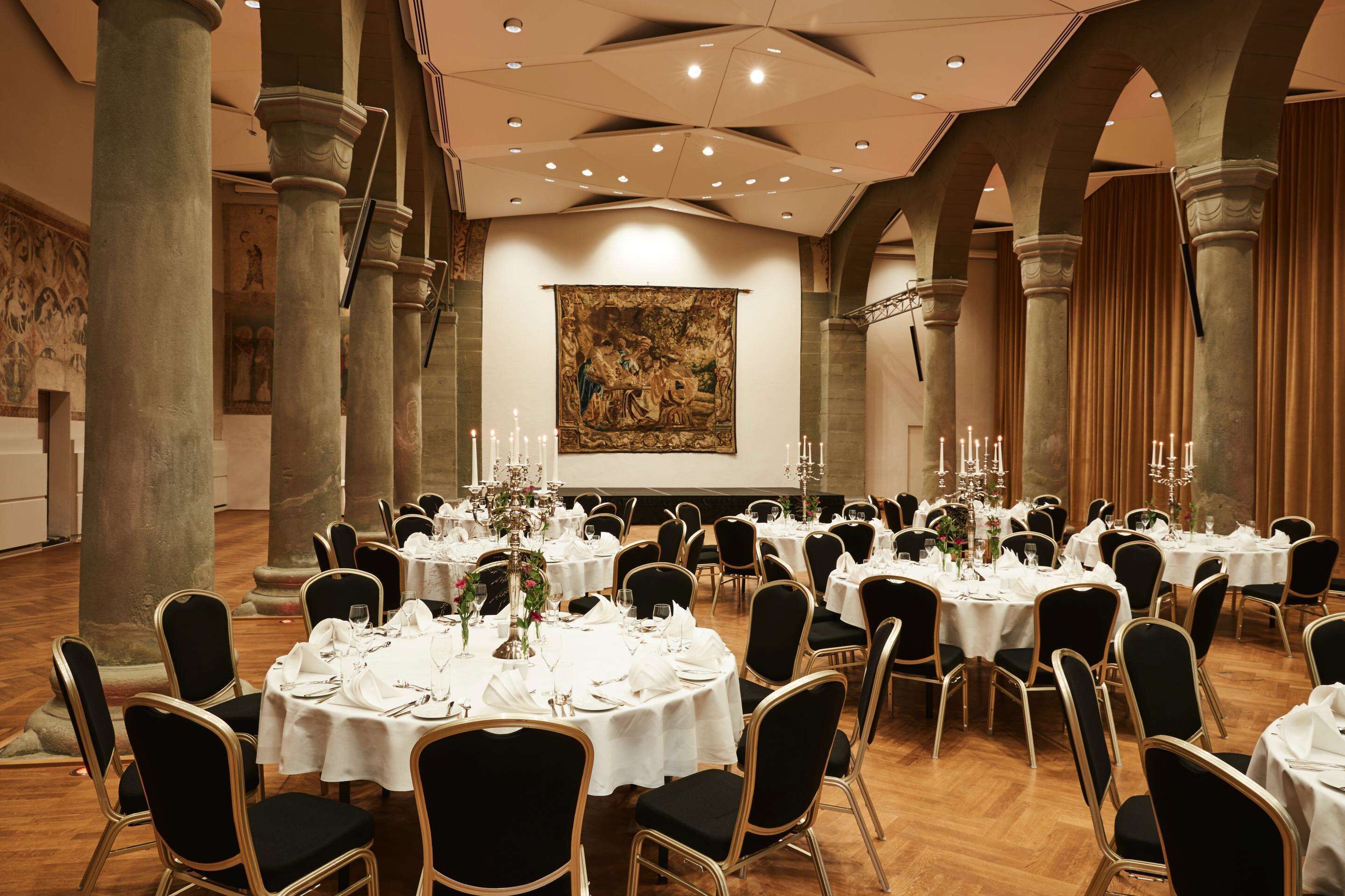 Un salón de banquetes con mesas redondas dispuestas para comer, con sillas negras y manteles blancos.