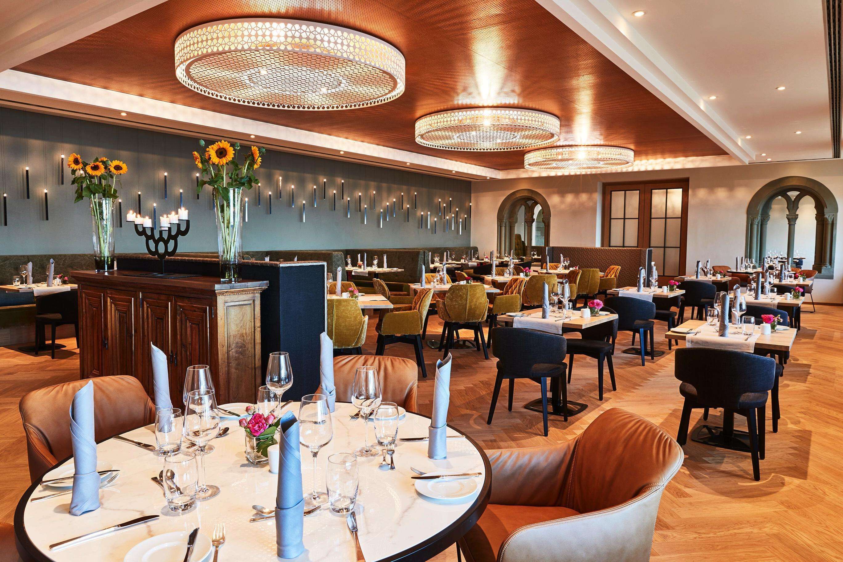 A restaurant with round tables, brown leather chairs, and chandeliers.