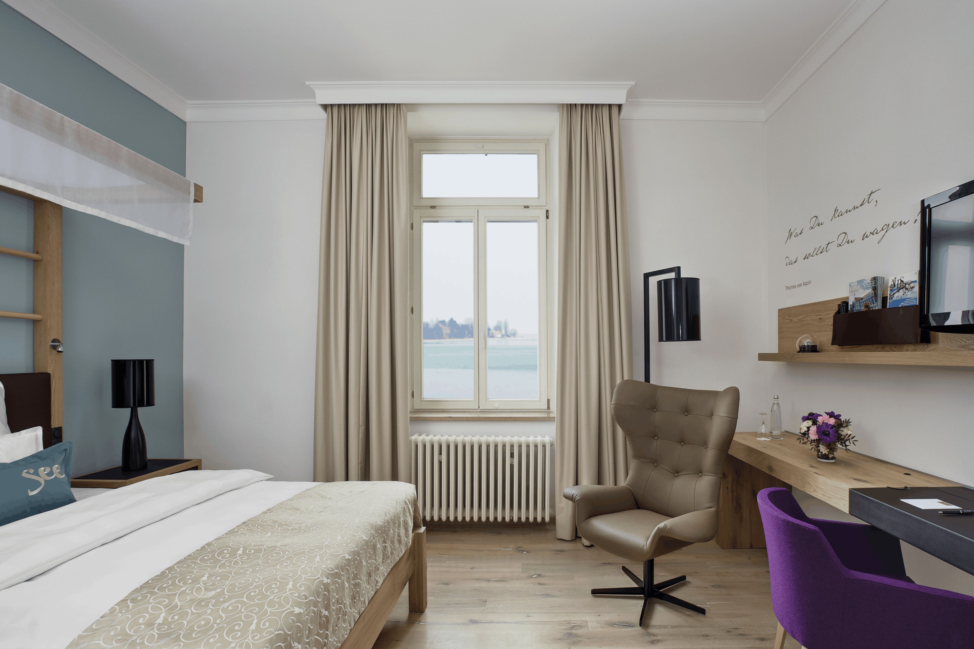 A hotel room with a bed, a desk, a chair, a radiator, and a window with curtains.