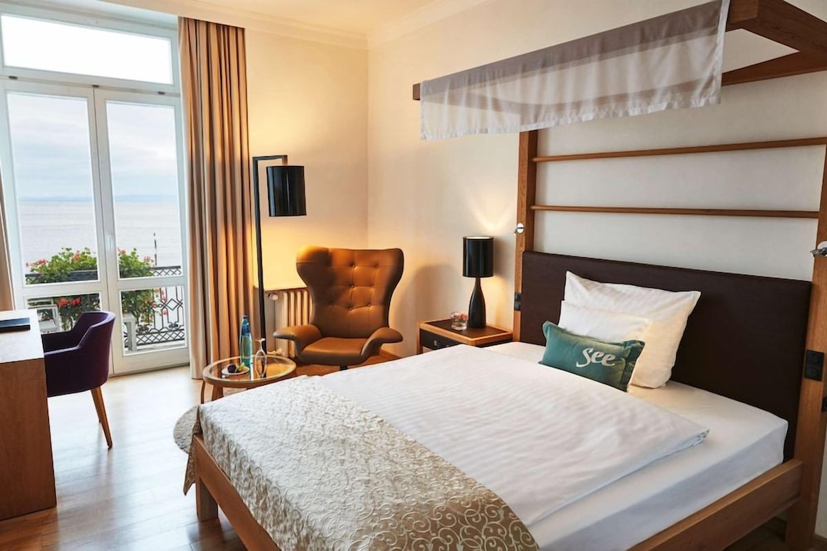 Habitación de hotel con cama, una silla, mesita de noche, lámpara y vista al mar.