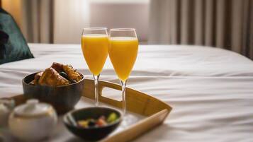 En välordnad frukostbricka med två glas apelsinjuice, en skål med frukt och en liten tekanna, placerad på en säng med utsikt över ett fönster och gardiner.