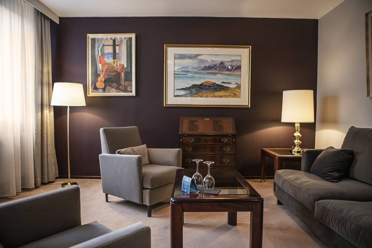 Een hotelkamer met een bank, fauteuil en een klein tafeltje met een lamp en twee wijnglazen. Twee schilderijen hangen aan de muur en er is een dressoir met lades.