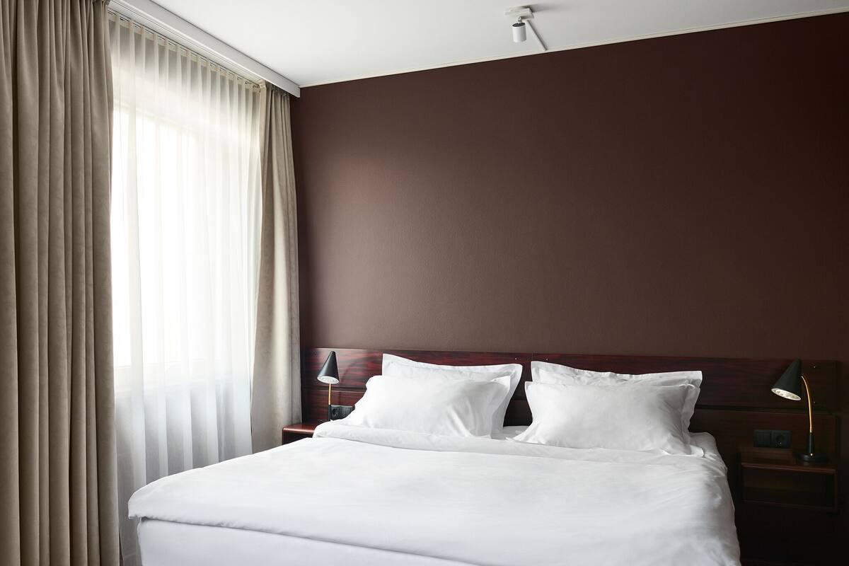 Een net opgemaakt bed met witte lakens in een kamer met bruine wanden en een groot raam met gordijnen.