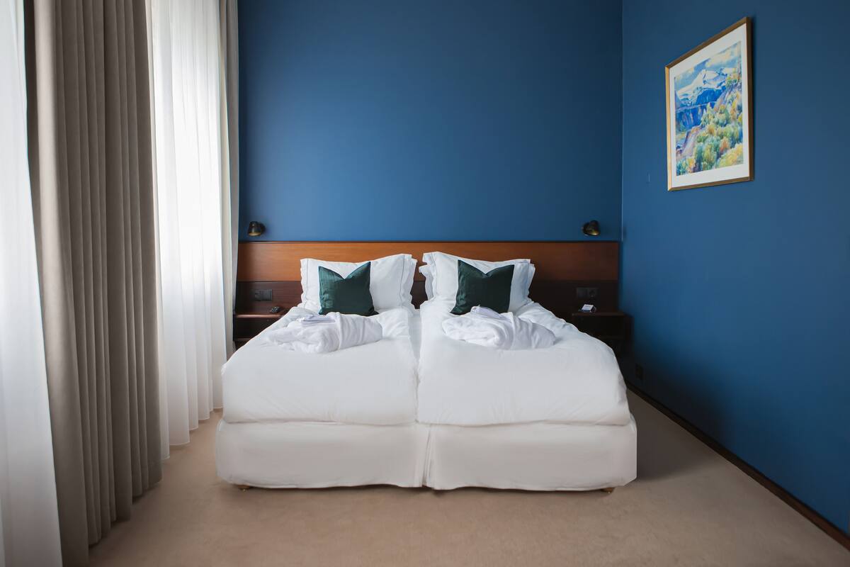 Een tweepersoonsbed met witte lakens en groene kussens, een houten hoofdbord, een nachtkastje met een lamp en een schilderij op de blauwe muur.