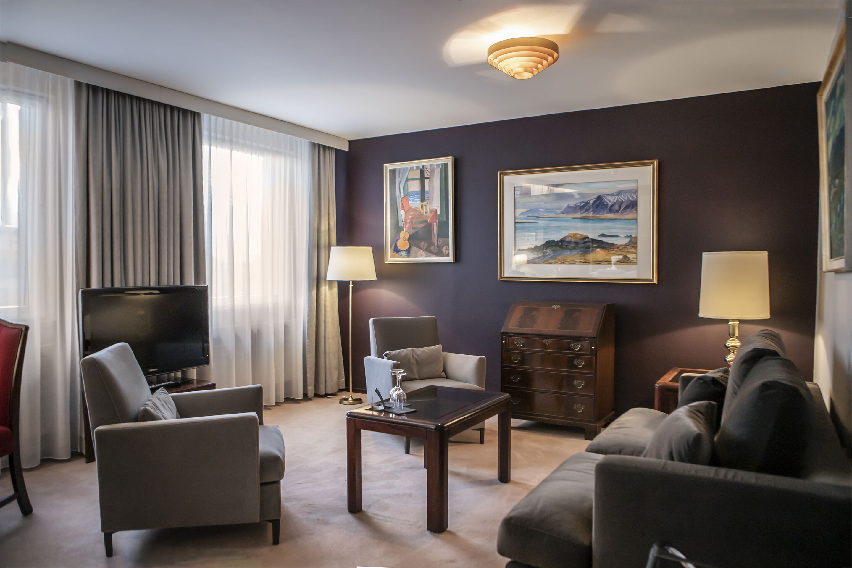 Habitación de hotel con televisión de pantalla plana, dos sillones, un sofá, una mesa de centro, un aparador y dos cuadros en la pared.