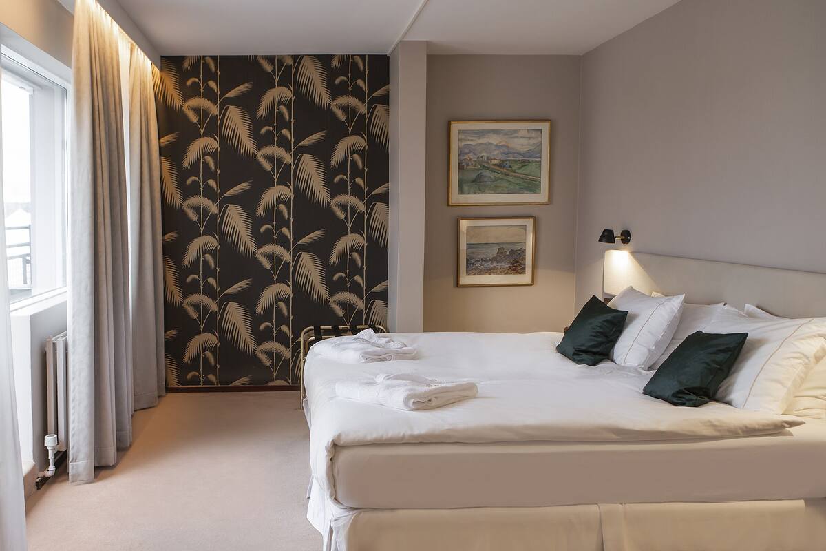 Een net opgemaakt bed met witte lakens en groene kussens, een radiator, een raam met gordijnen, en twee ingelijste foto's aan de muur.