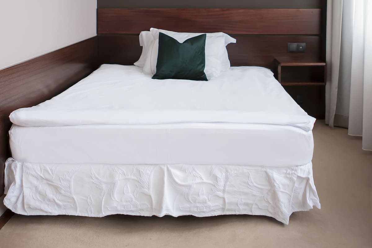 Een netjes opgemaakt bed met witte beddengoed en een groen kussen, een lijst met kunstwerk aan de muur, en een nachtkastje met een lamp.