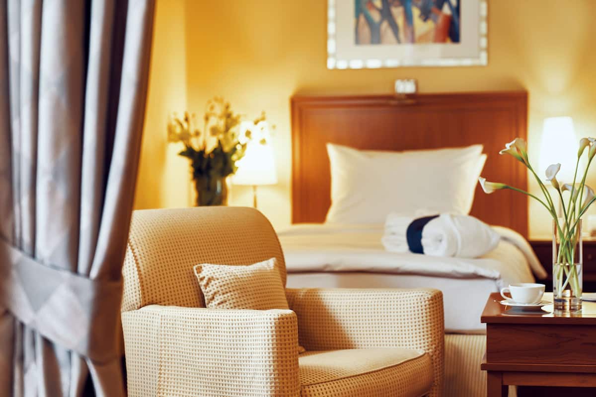 A hotel room with a bed, a chair, a bedside table with a cup, and a vase with flowers.