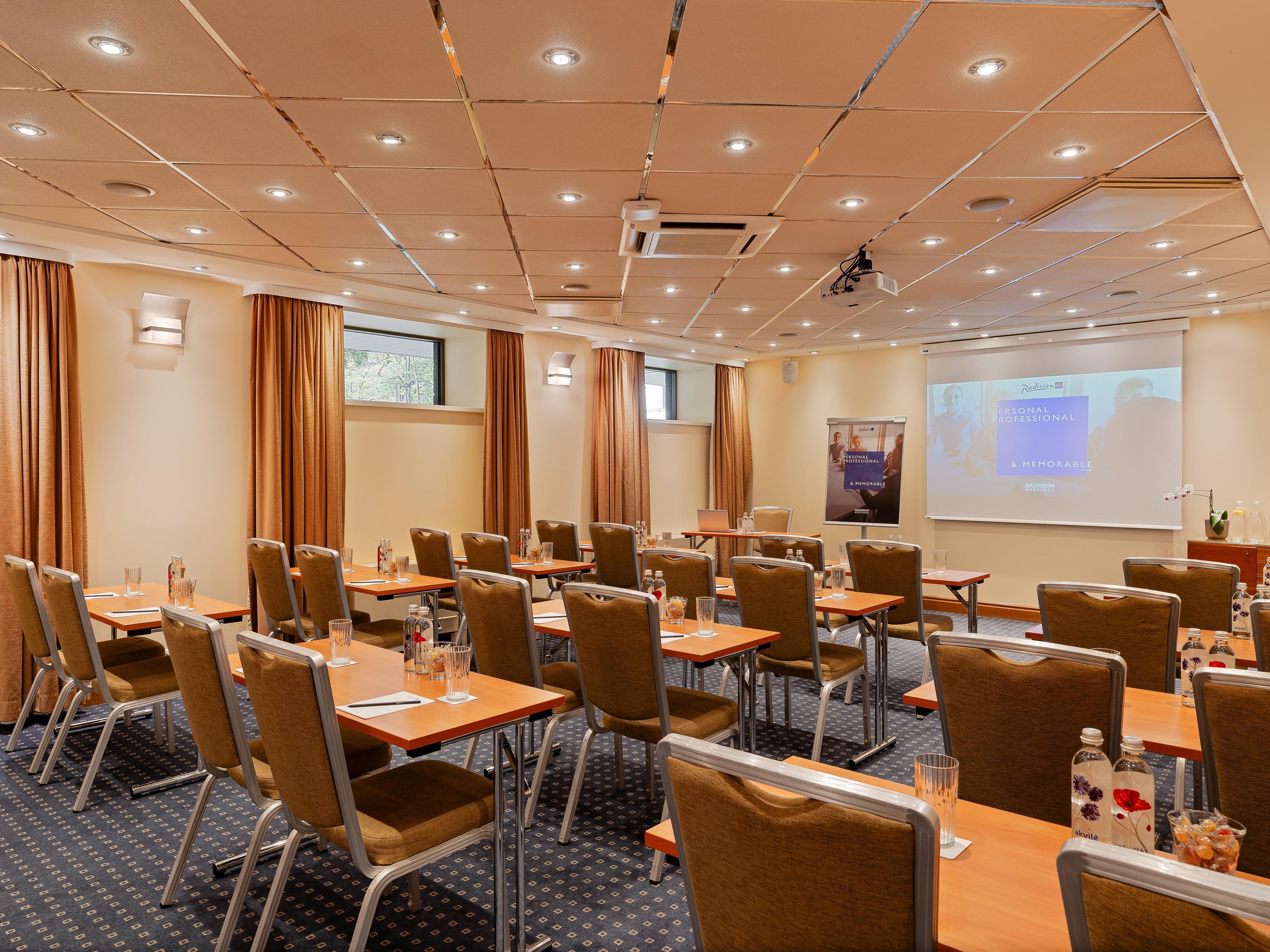 Un salón de conferencias con mesas, sillas, una pantalla de proyector y una pantalla para presentaciones.