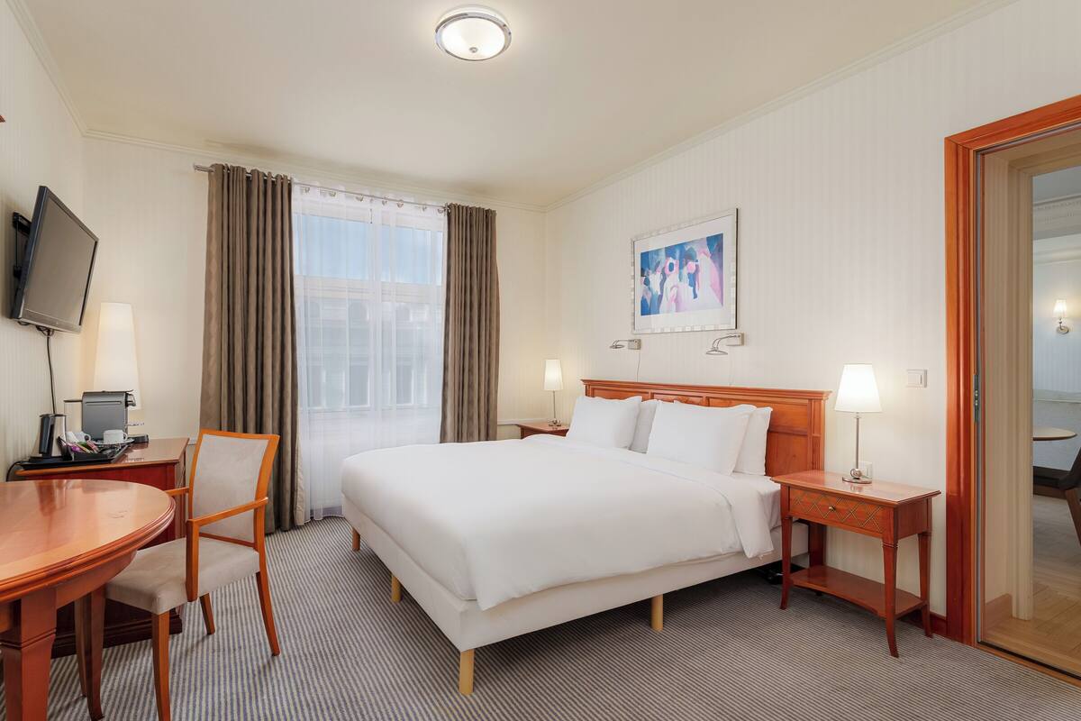 A hotel room with a bed, bedside tables, a desk with a chair, a TV, and a window with curtains.