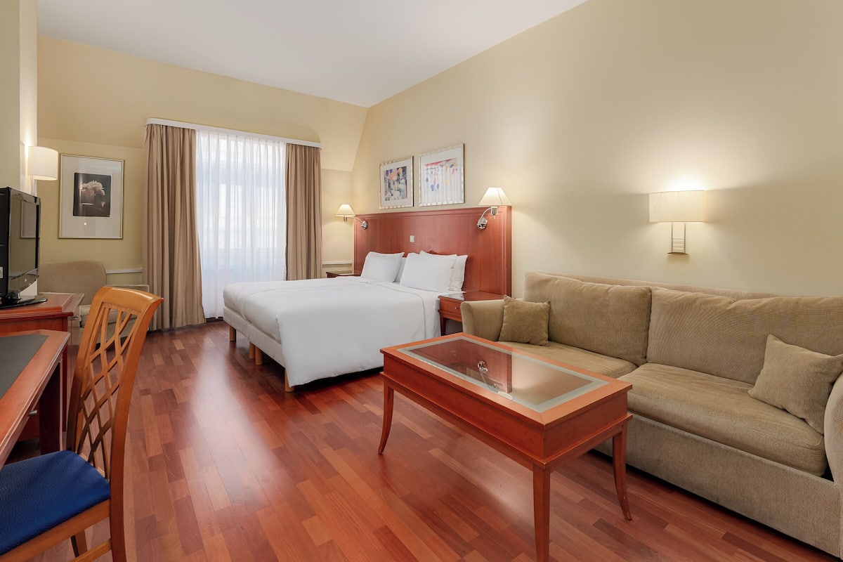 A hotel room with a bed, a sofa, a coffee table, a desk with a chair, and a TV.