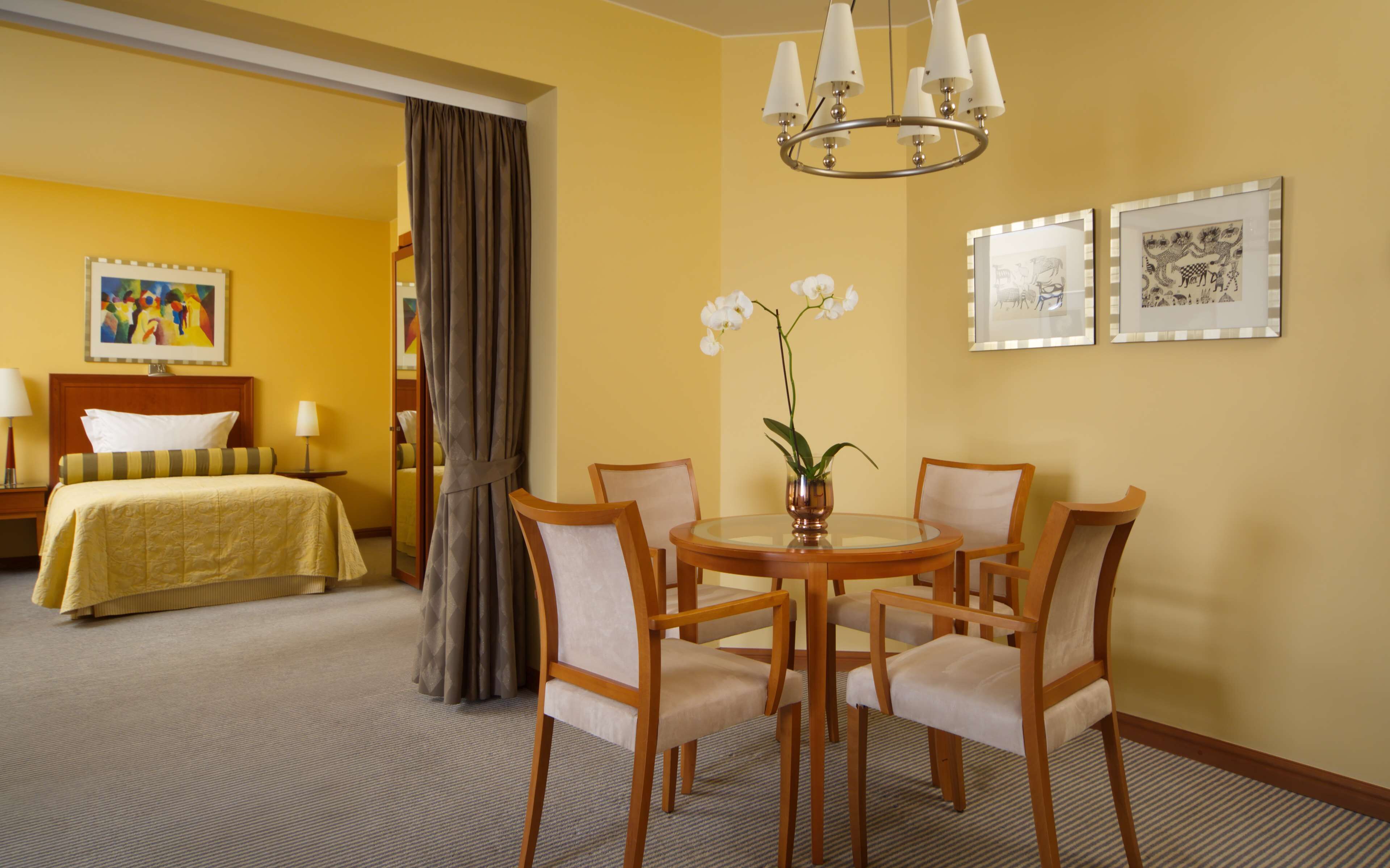 A hotel room with a bed, a dining table with chairs, a vase with flowers, a chandelier, and framed pictures on the wall.