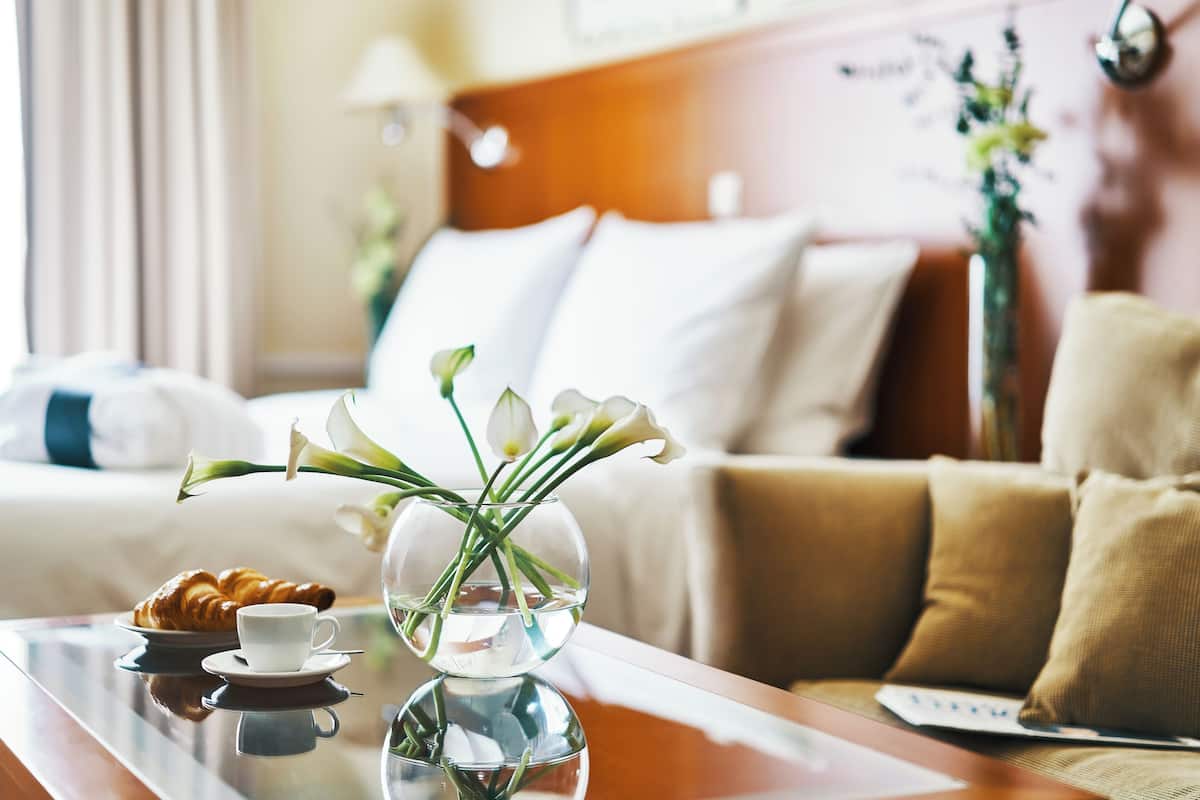 A hotel room with a bed, a bedside table with a croissant and a cup, and a vase with flowers.