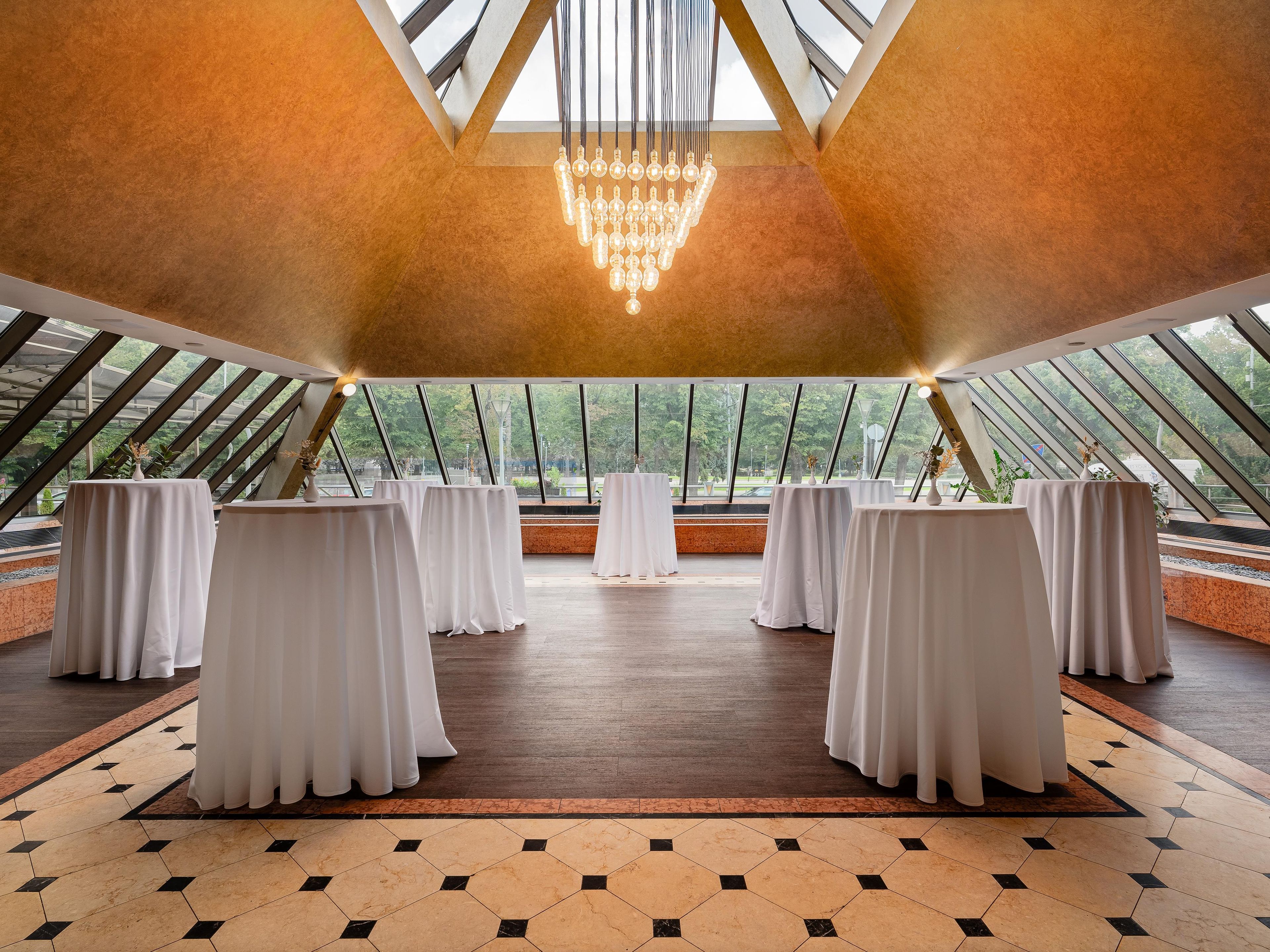 A spacious room with a high ceiling, large windows, and a chandelier. Tables are arranged for seating.