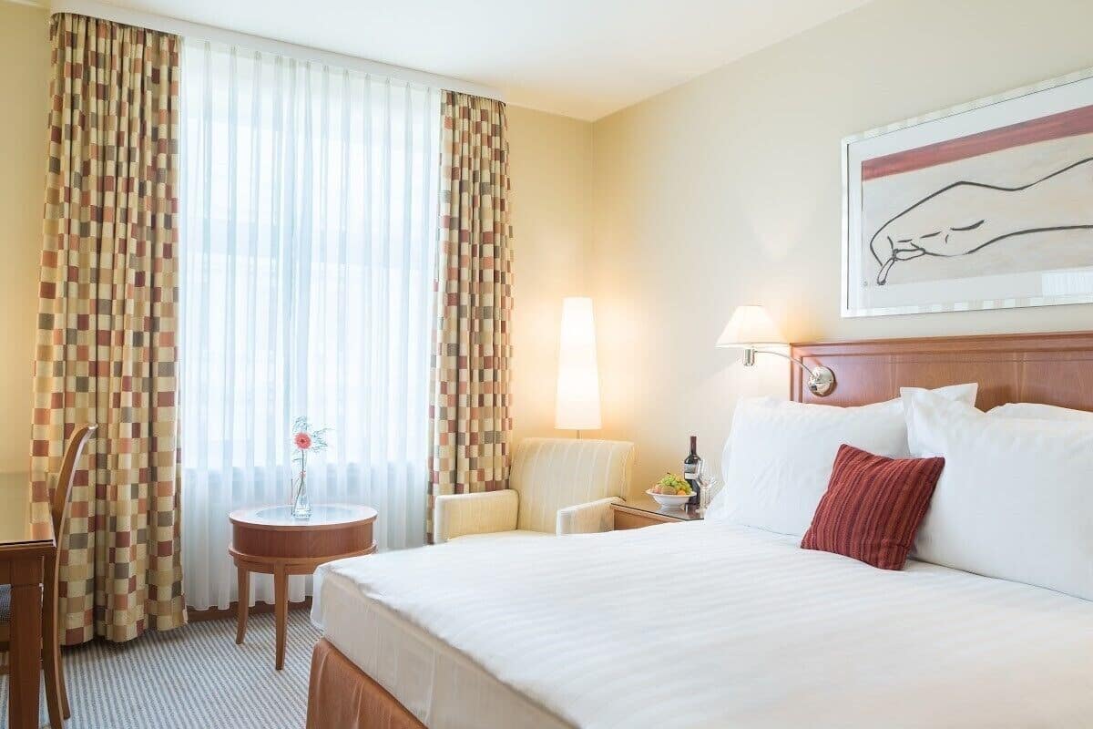 A hotel room with a large bed, a bedside table with a lamp, a chair, and a small table with a vase of flowers.