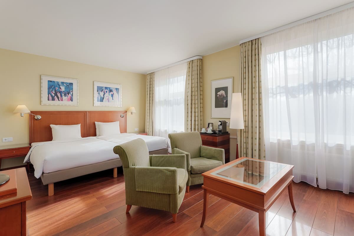 A hotel room with a bed, two armchairs, a glass-top table, wooden flooring, and curtains.