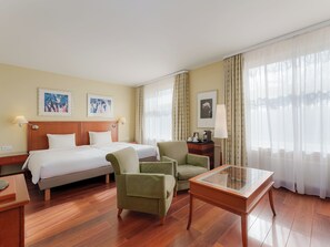 A hotel room with a bed, two armchairs, a glass-top table, wooden flooring, and curtains.