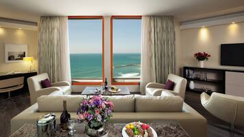 A modern hotel room with a large window offering a sea view, a sofa set, a dining table with a vase of flowers, and a flat-screen TV on the wall.