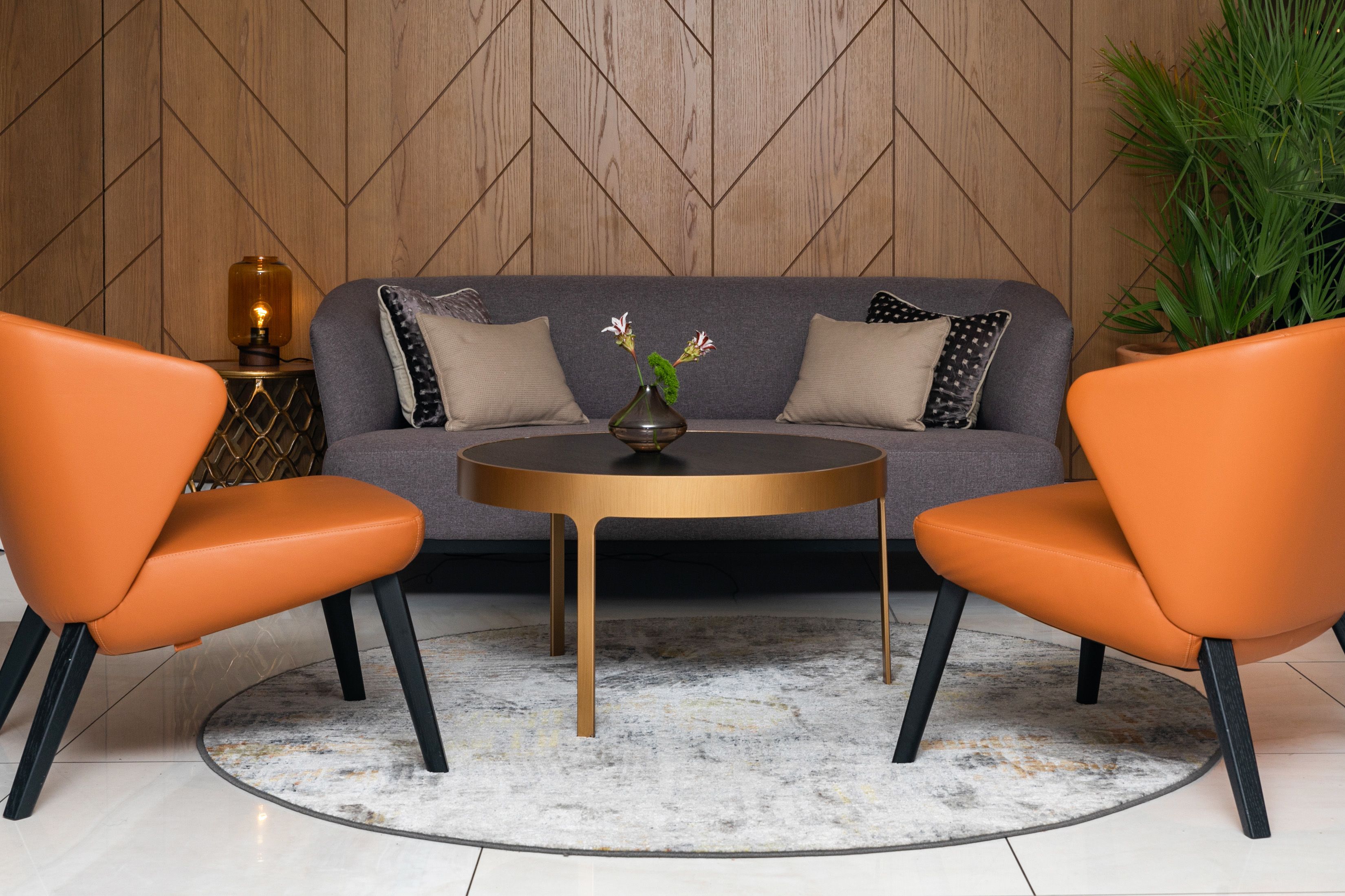 A modern living room with a sofa, two orange chairs, a round table, and a decorative vase.