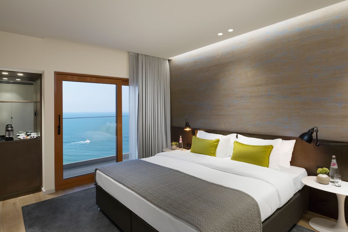 A modern hotel room with a large bed, a view of the ocean, a small table with a lamp, and a minibar.