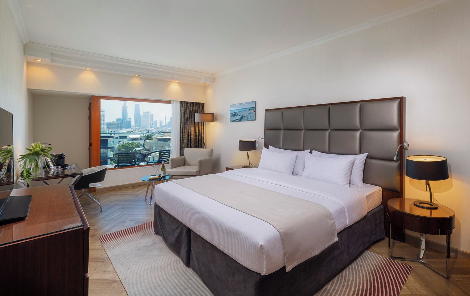 A hotel room with a large bed, a desk, a chair, a small table with a bottle and glasses, and a view of the cityscape.