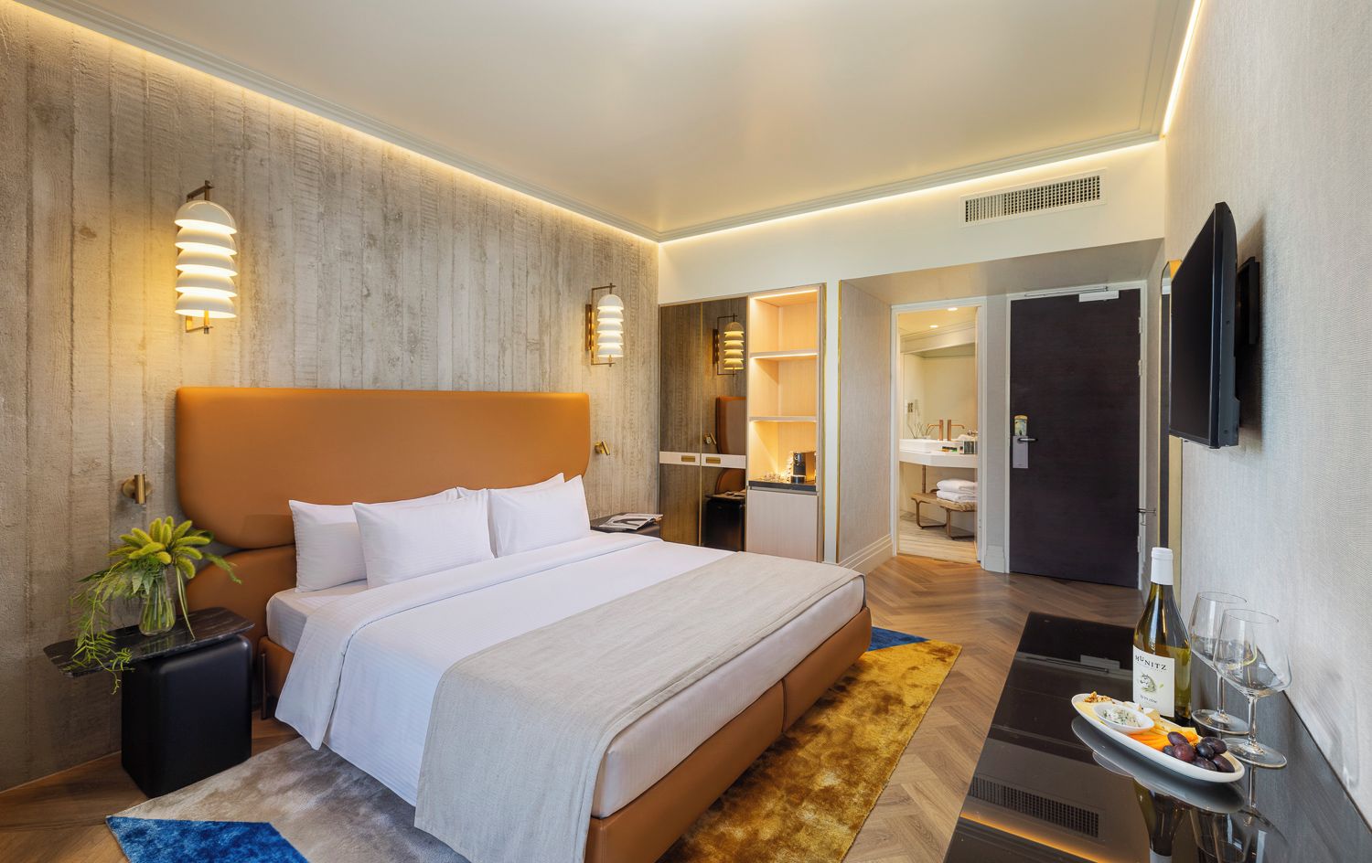 Ein modernes Hotelzimmer mit einem großen Bett, Nachttischen, einem kleinen Tisch, einer Topfpflanze, einer Minibar, einem Badezimmer und einem Fernseher.
