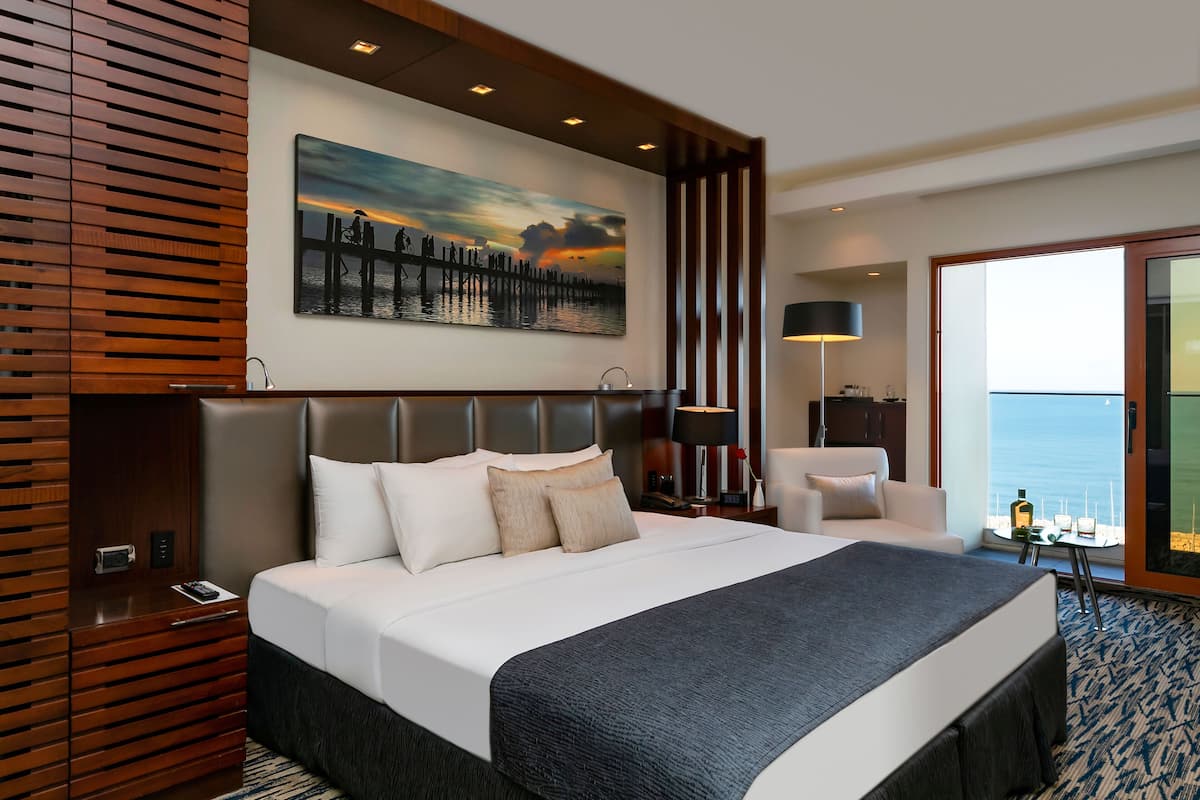 A modern hotel room with a large bed, a painting of a sunset over water, a view of the ocean through a glass door, and a wooden panel wall.