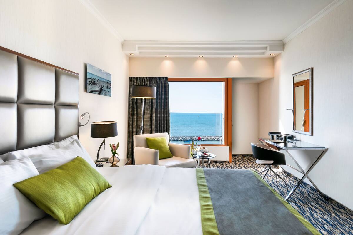 A hotel room with a bed, a desk, a chair, a lamp, and a view of the sea.