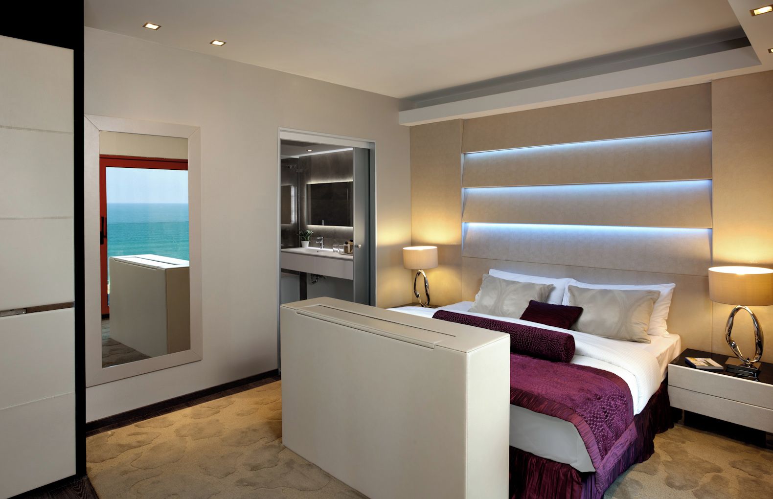 A modern hotel room with a bed, bedside tables, a mirror, and a view of the sea.