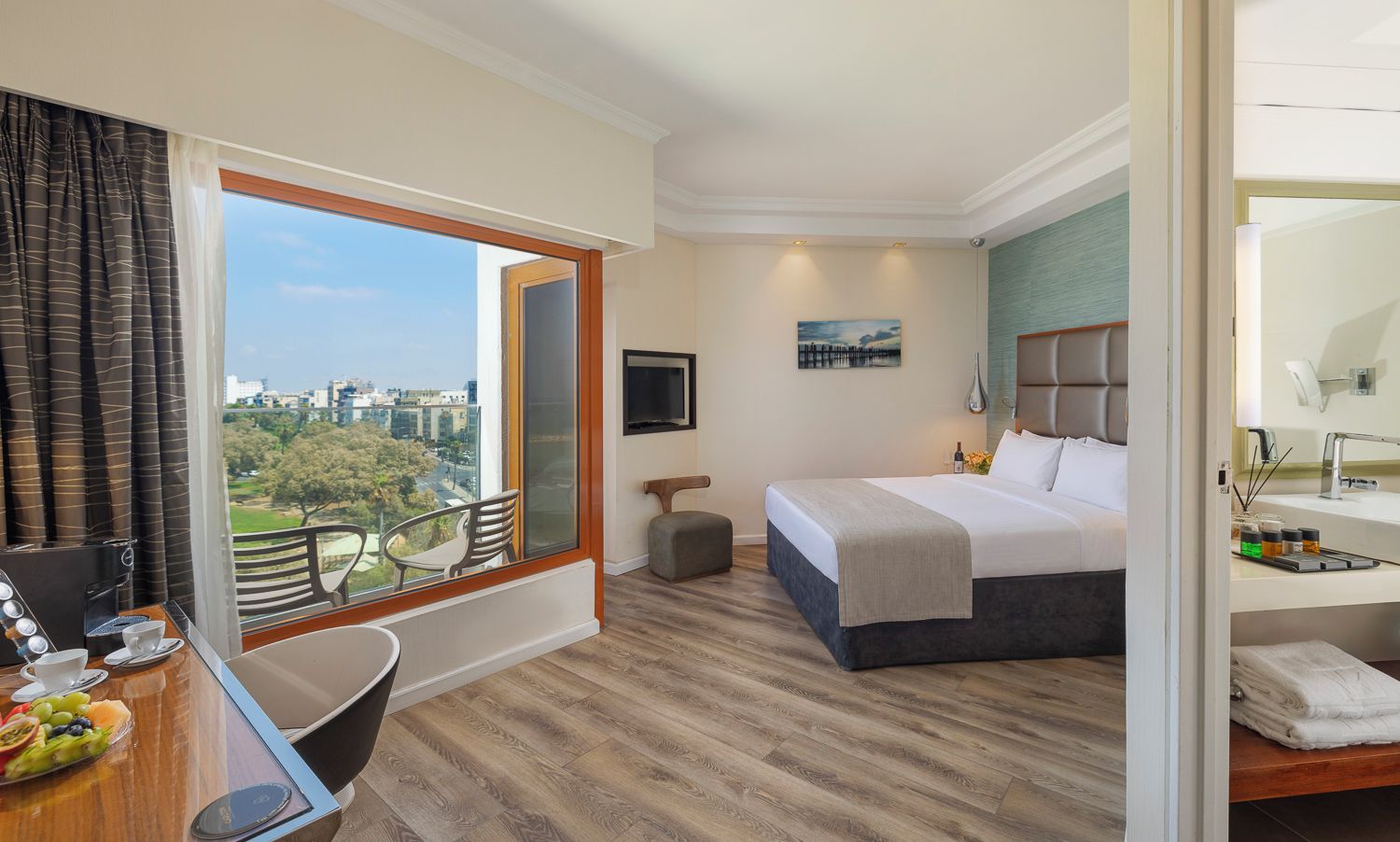 A hotel room with a bed, a desk with a chair, a small table with a fruit bowl, a window with curtains, and a city view.