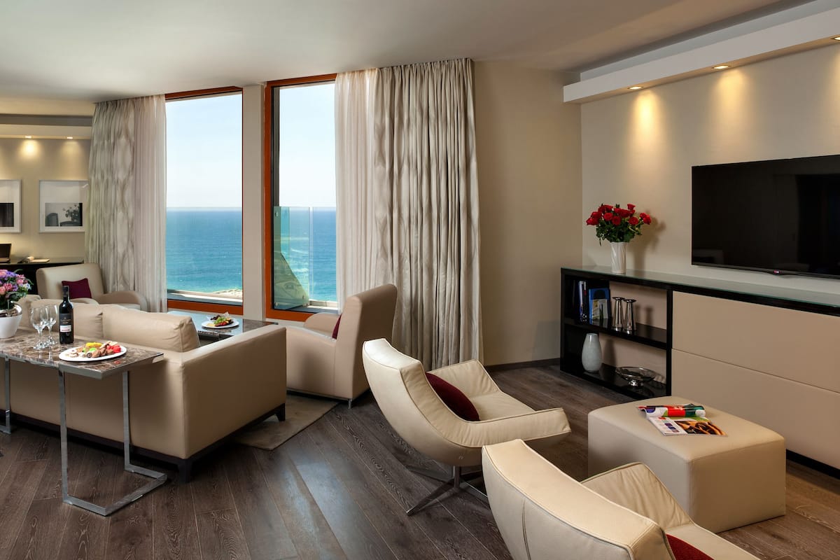 A modern hotel room with a dining area, a sofa, a TV, and a view of the ocean.