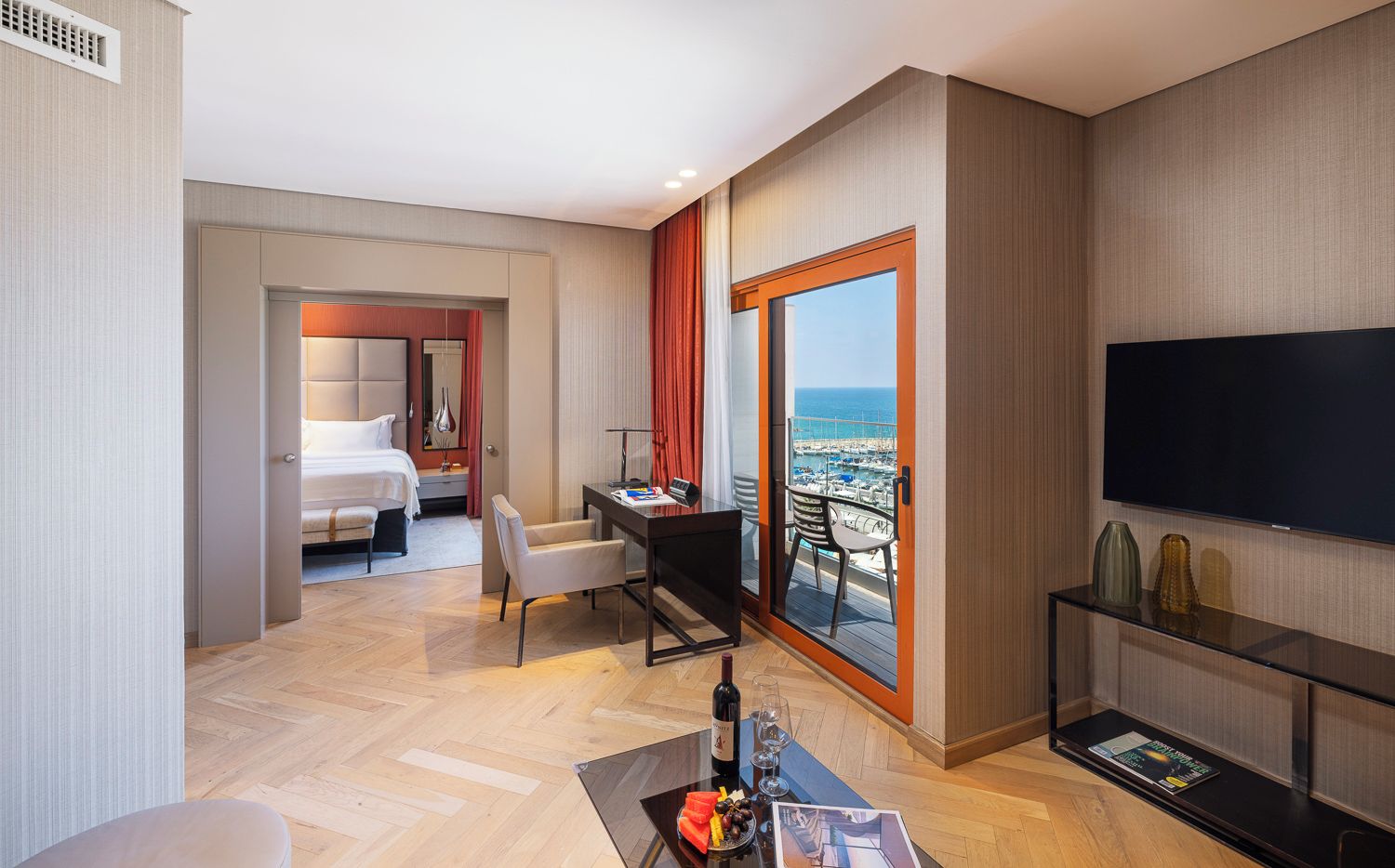 A hotel room with a bed, a desk, a TV, and a balcony with a view of the sea.