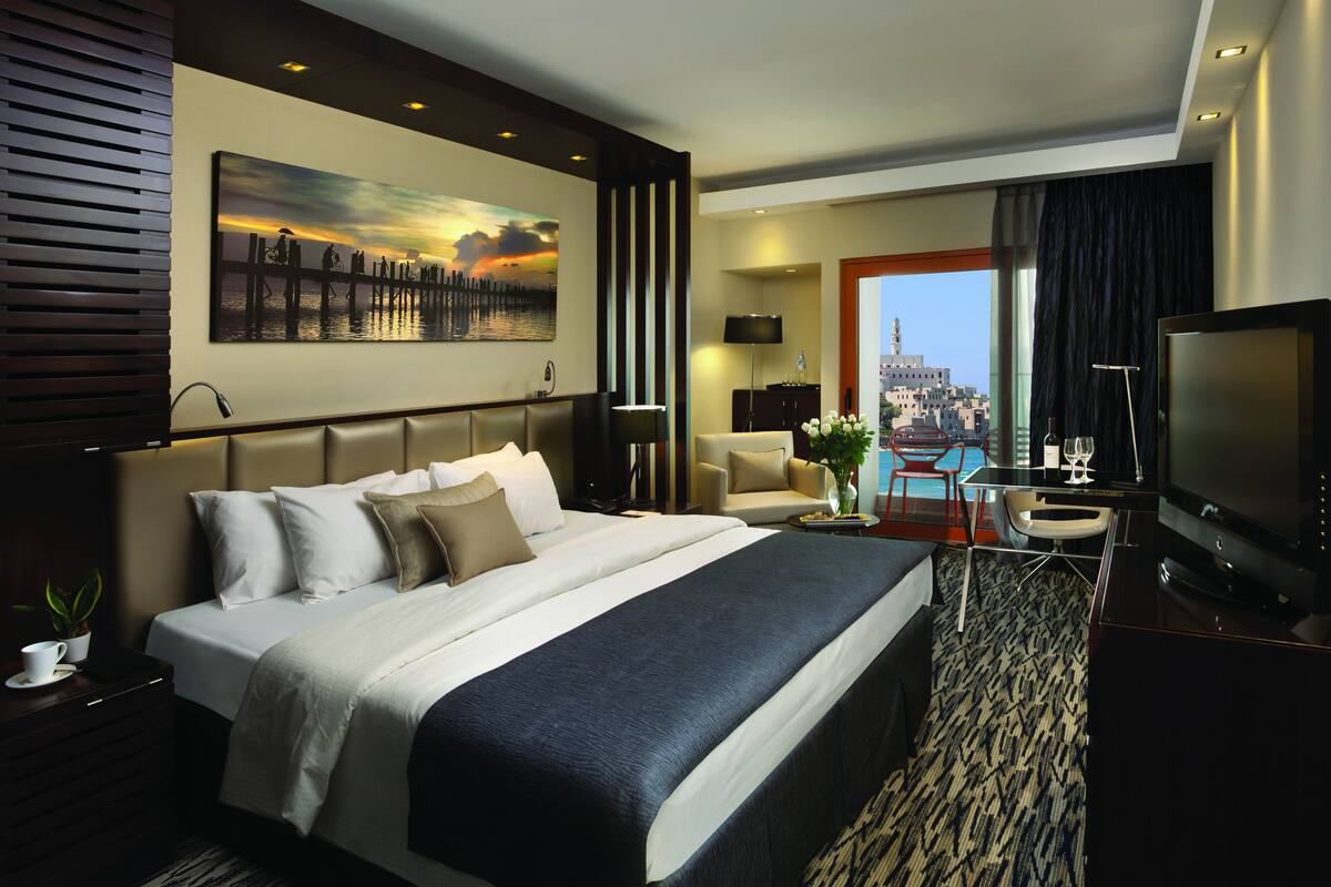 A modern hotel room with a large bed, a desk with a chair, a TV, and a view of the cityscape.