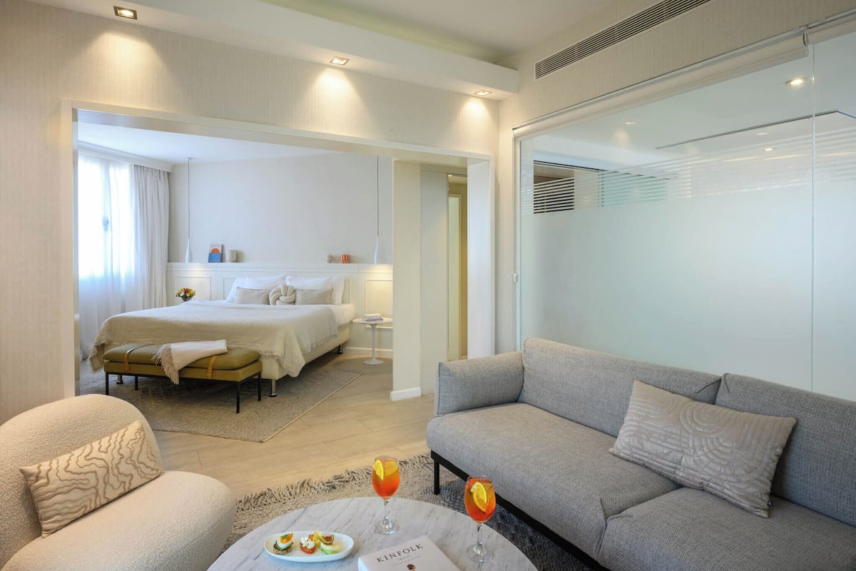 A modern hotel room with a bed, a sofa, a coffee table with a plate of food and drinks, and a view of the hallway.