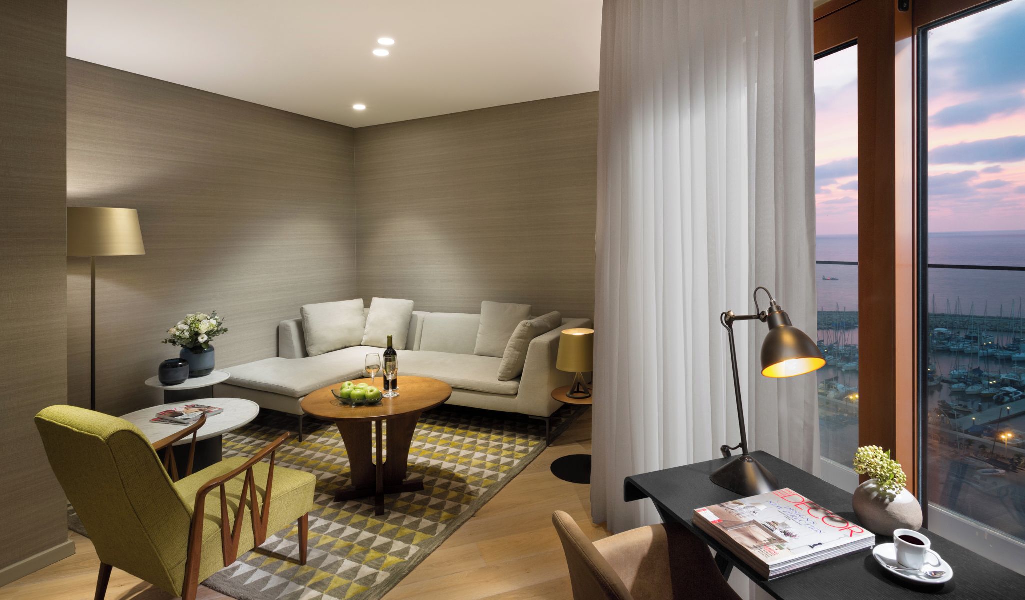 Une chambre d’hôtel moderne avec un canapé, une table ronde, une table basse, un bureau avec une lampe et une vue sur le port de plaisance.