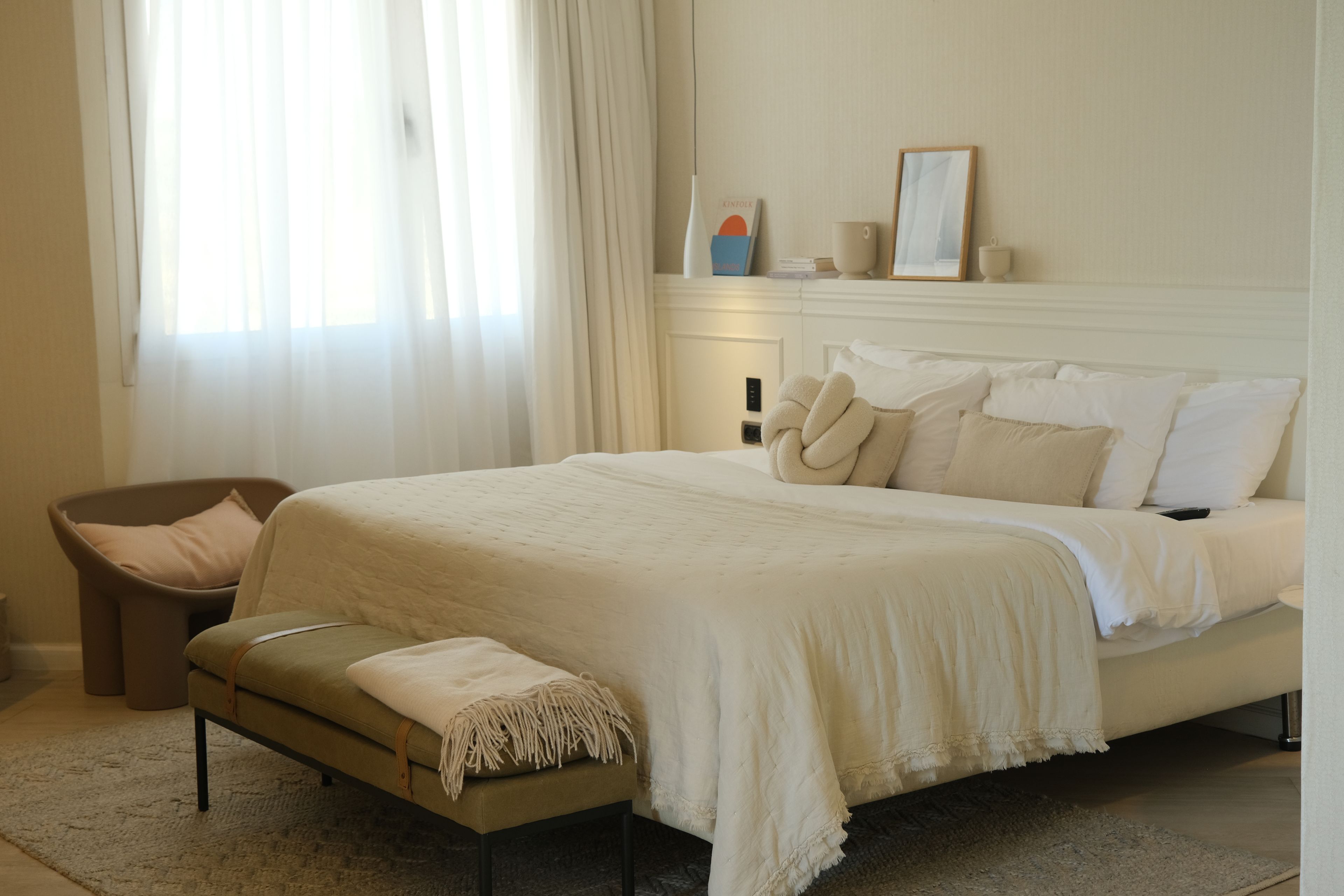A bedroom with a bed, a bench, a chair, a bedside table, and a window with curtains.