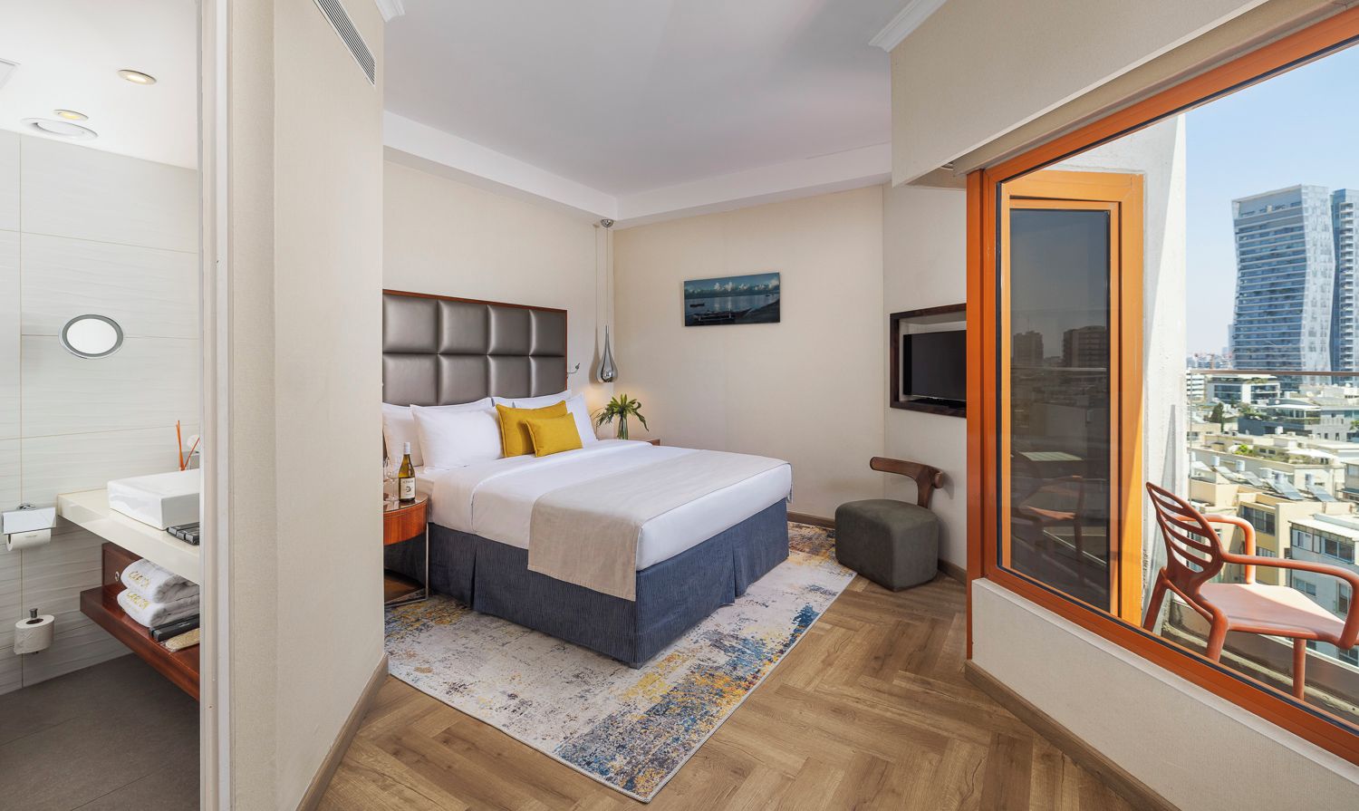 A modern hotel room with a large bed, a TV, a balcony with a chair, and a city view.