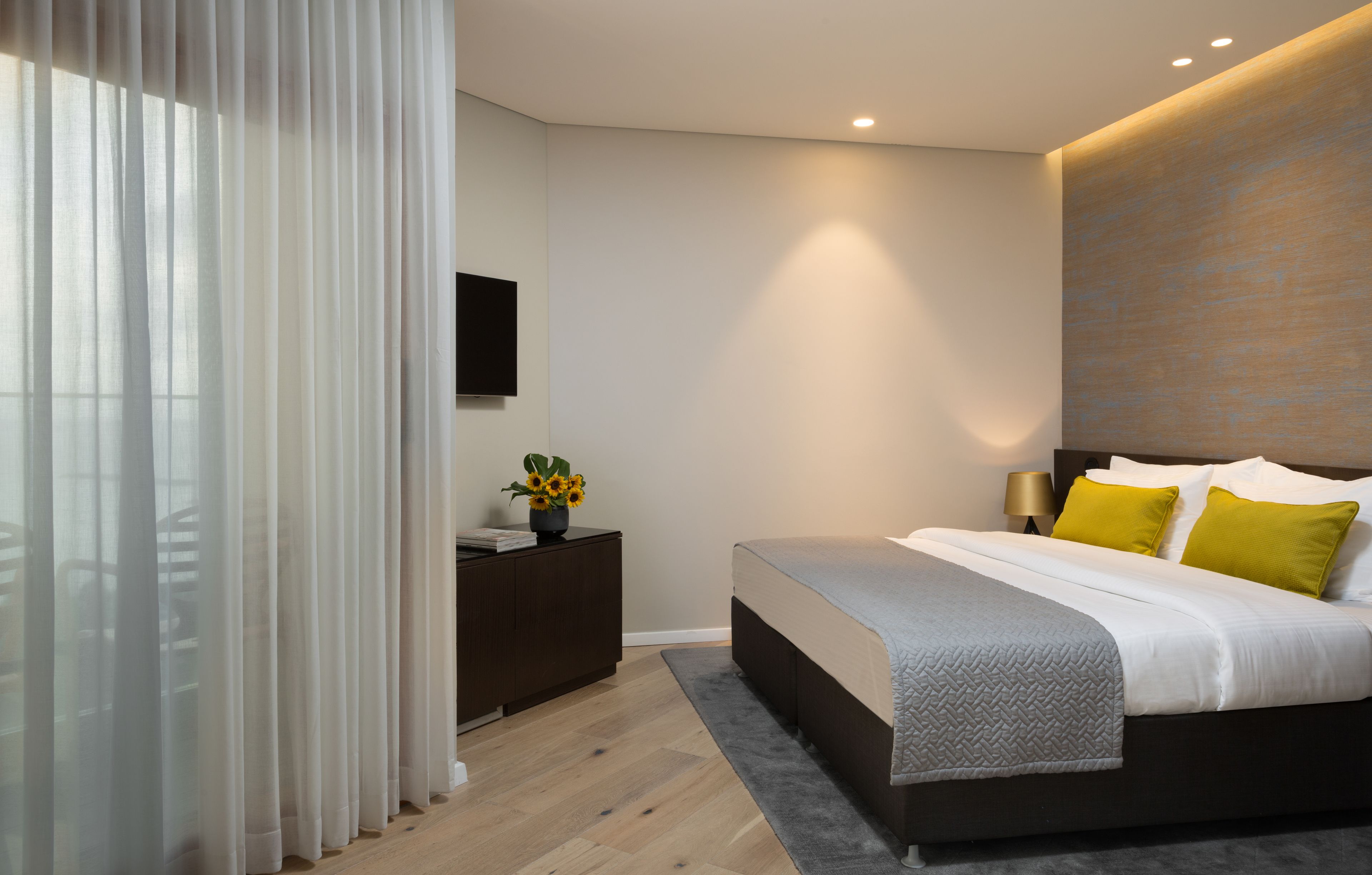 Een moderne slaapkamer met een groot bed, nachtkastje, televisie en raam met dunne gordijnen.