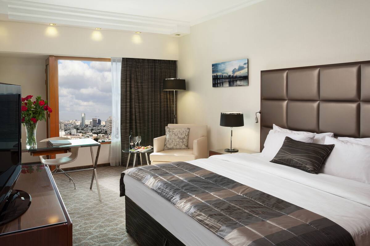 A hotel room with a large bed, a desk with a chair, a TV, a lamp, and a view of the city.