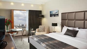 A hotel room with a large bed, a desk with a chair, a TV, a lamp, and a view of the city.