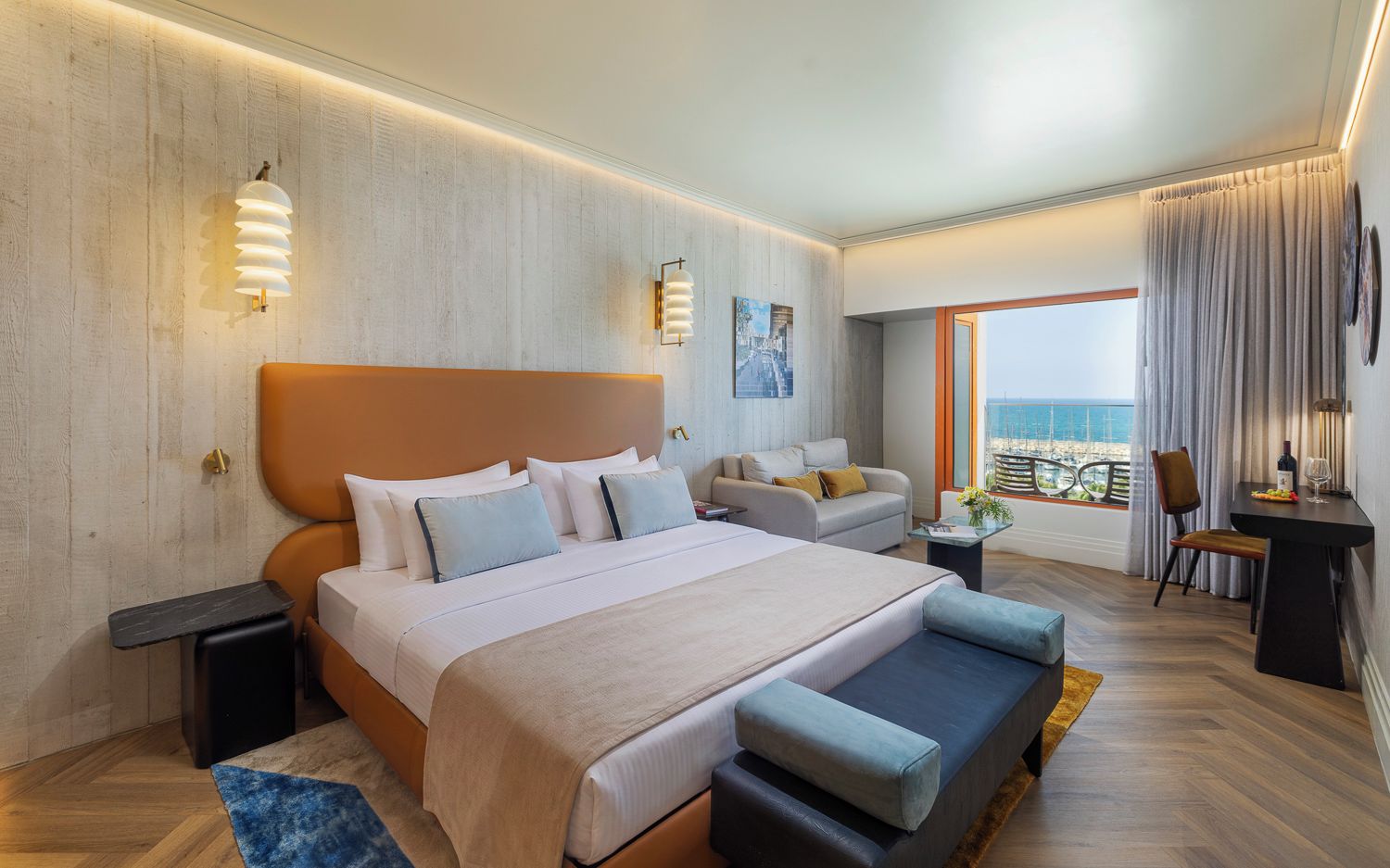 A modern hotel room with a large bed, a seating area, a dining table, and a view of the sea.