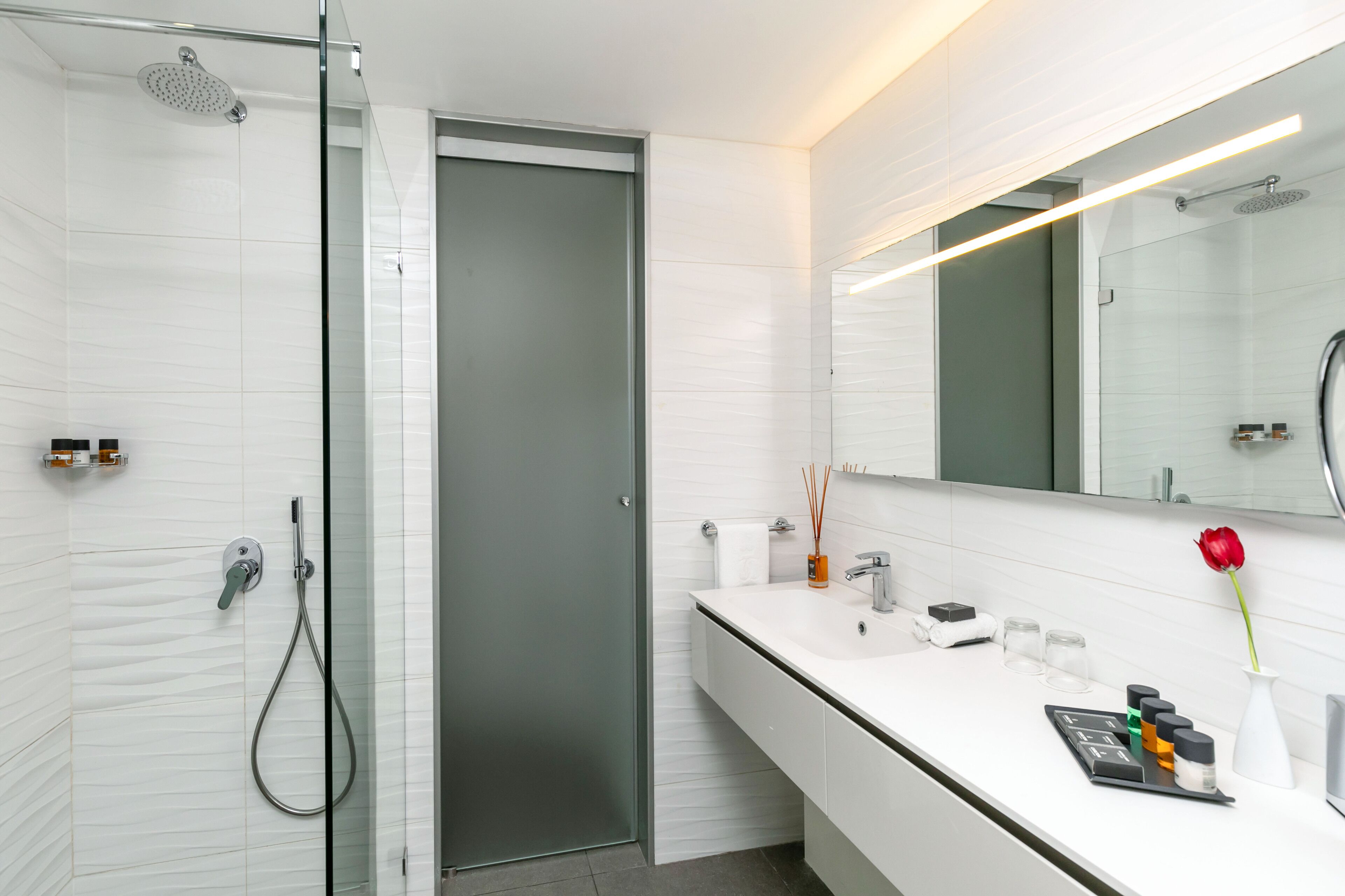 A modern bathroom with a glass shower enclosure, a white vanity with a mirror, and a red rose on the counter.