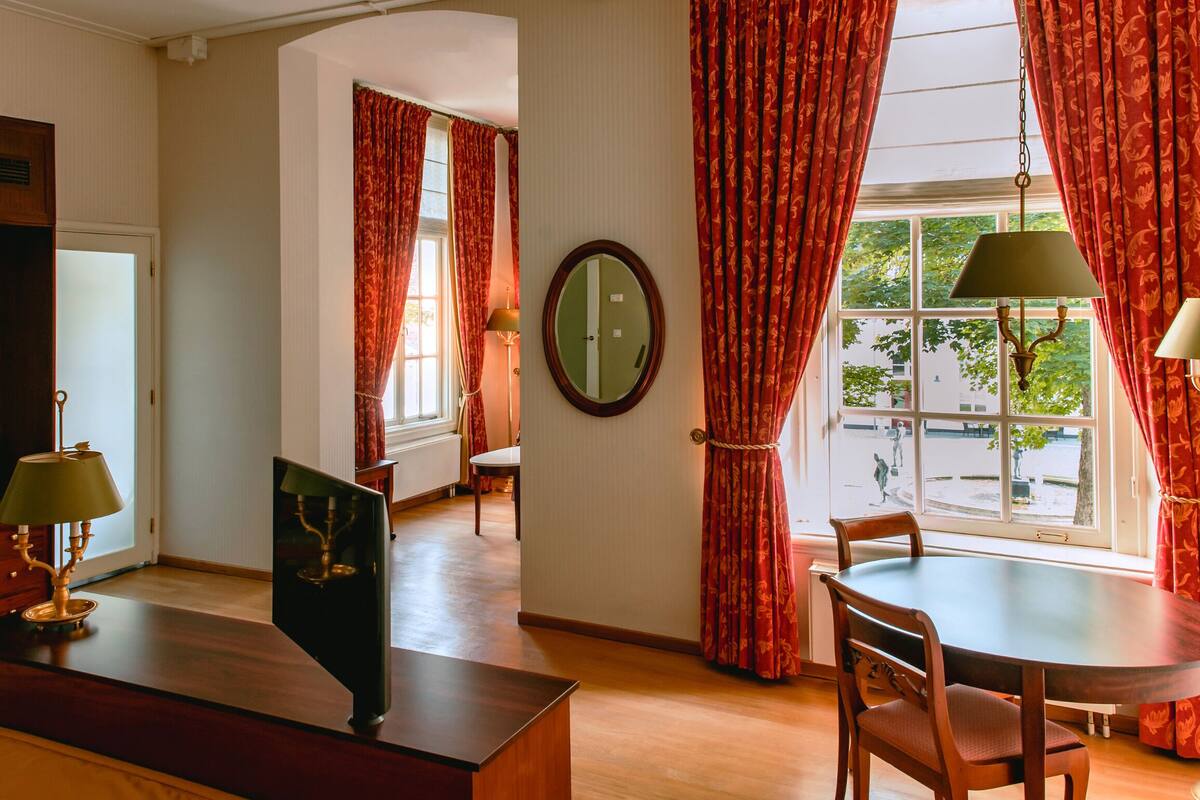 A room with a desk, chairs, a dining table, and a large window with red curtains.