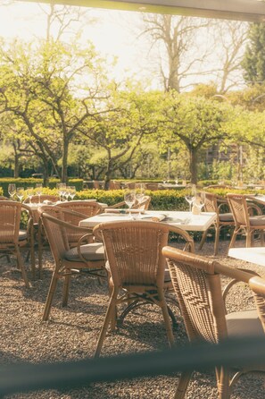 An outdoor dining area with wicker chairs and tables, surrounded by greenery and trees. - Grand Hotel Karel V, WorldHotels Luxury (Utrecht)