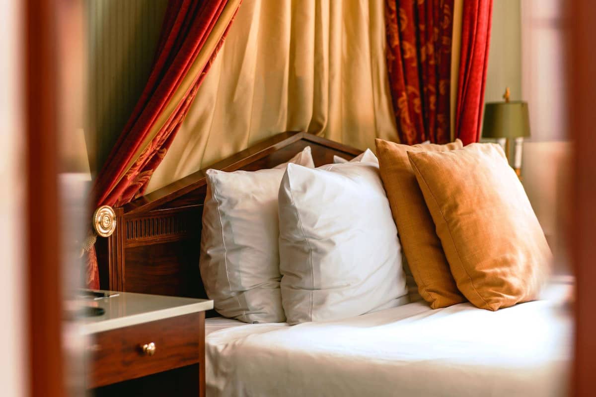 A neatly made bed with white and mustard pillows, a wooden headboard, and red curtains.
