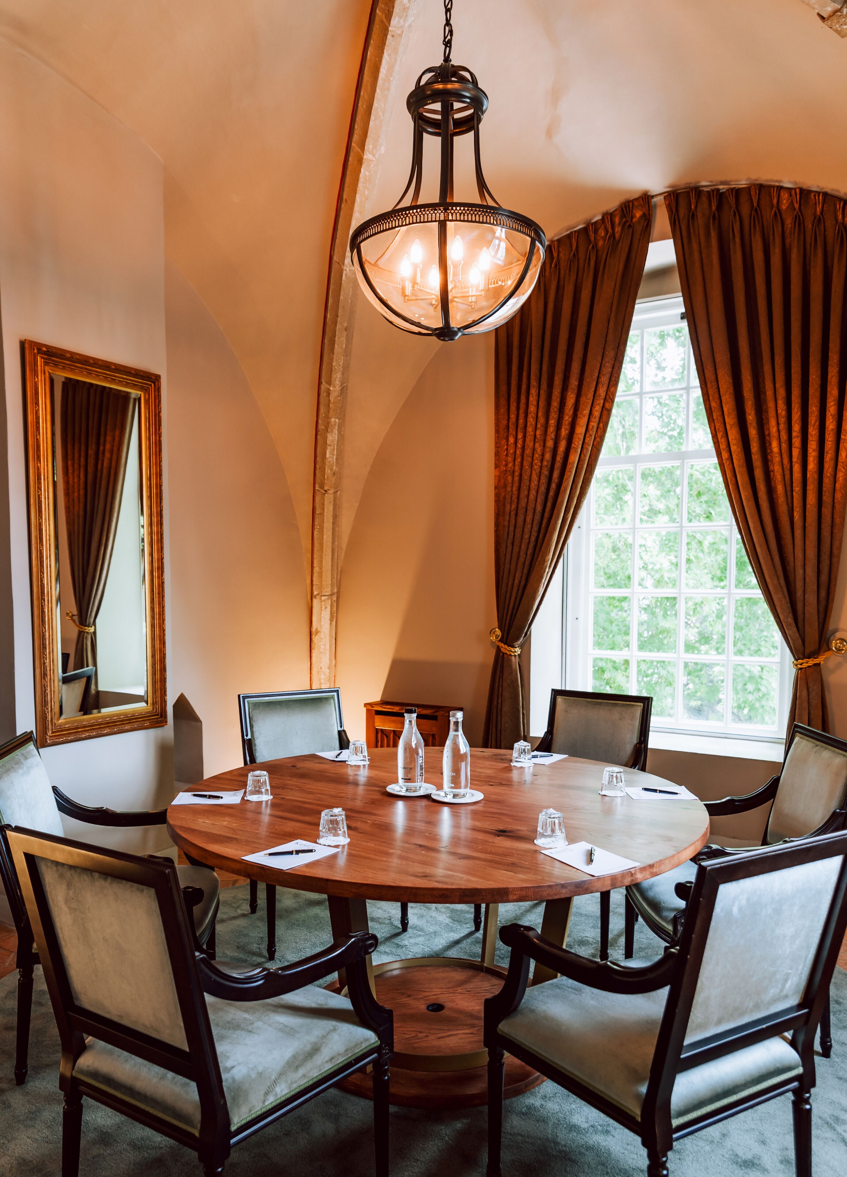 Een vergaderruimte met een ronde tafel, stoelen, een kroonluchter, een grote spiegel en een raam met gordijnen.