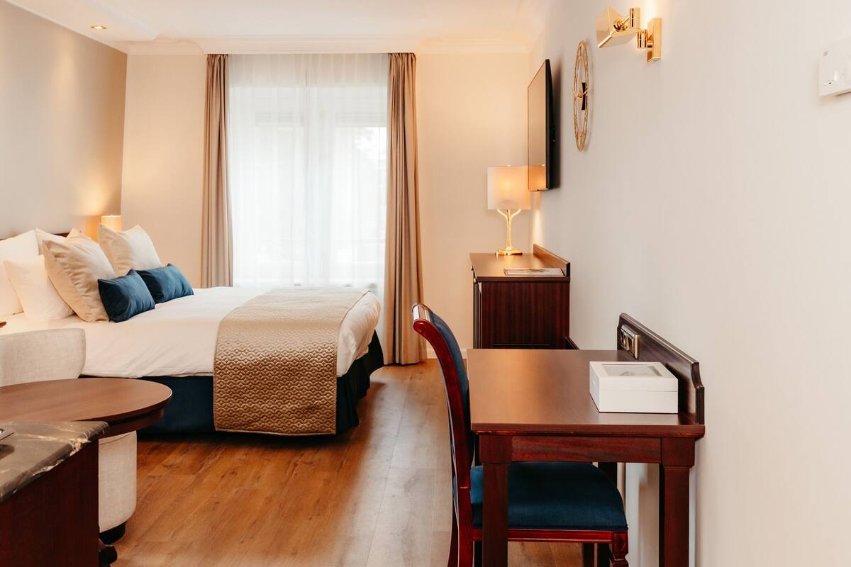 A hotel room with a bed, a desk, a chair, a lamp, and a TV mounted on the wall.