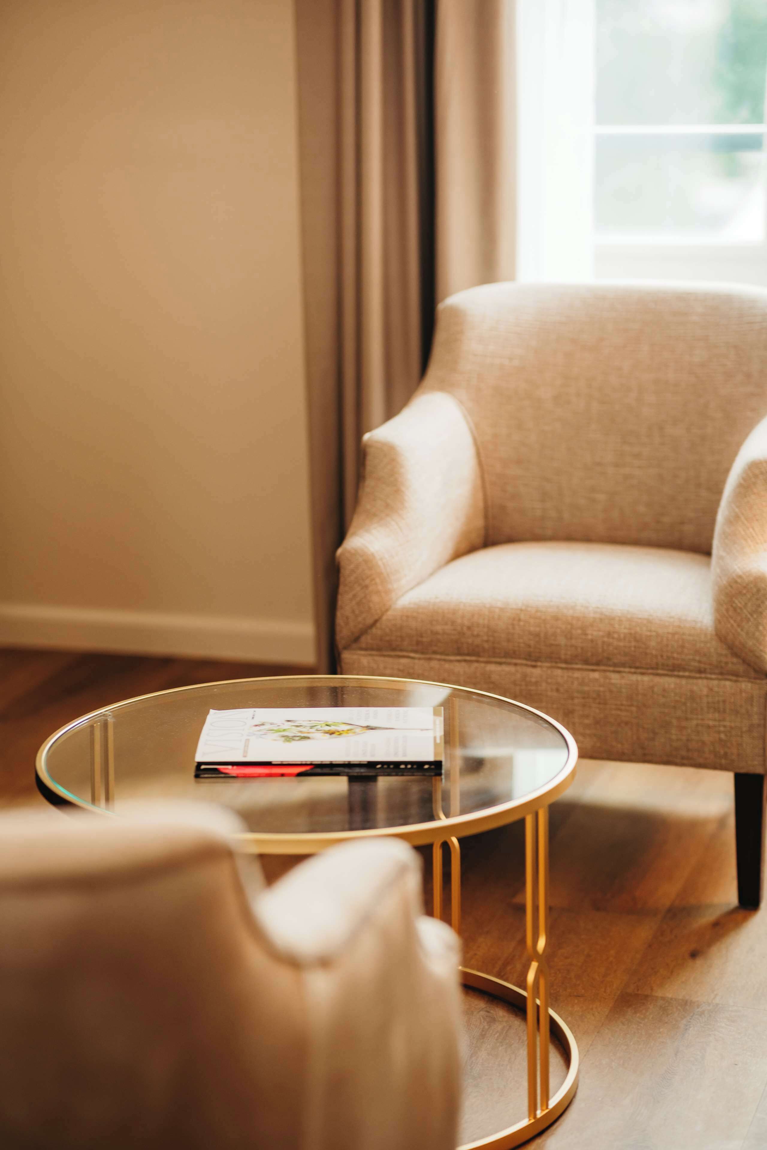 A living room with a beige armchair, a glass coffee table with a magazine, and a wooden floor.
